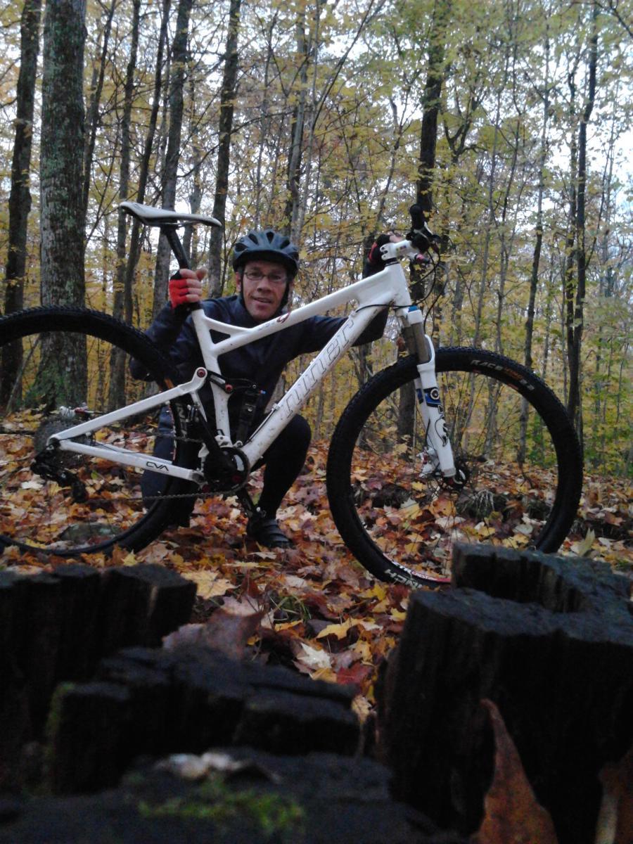 Niner Jet 9: A person wearing a helmet and gloves poses next to a white mountain bike in a forest during autumn. The ground is covered with colorful fallen leaves, and trees with yellow leaves are visible in the background.
