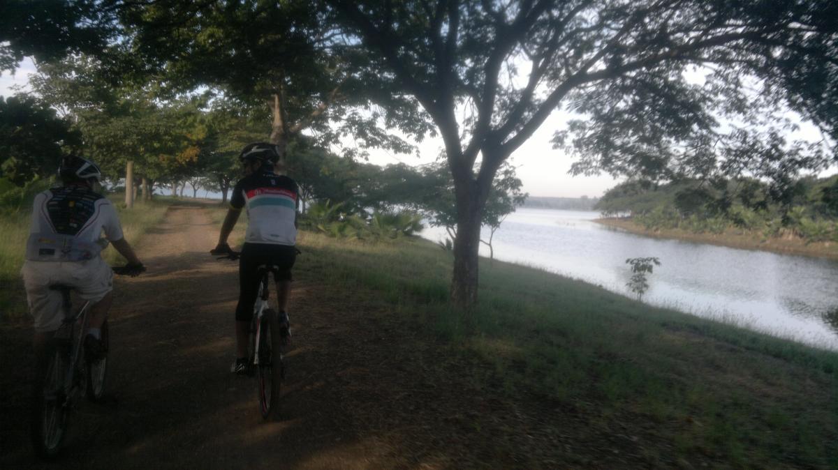 Two cyclists riding along a dirt path next to a river, surrounded by trees and greenery. The scene captures a serene outdoor environment with clear skies and natural beauty. La Primavera mountain bike trail.