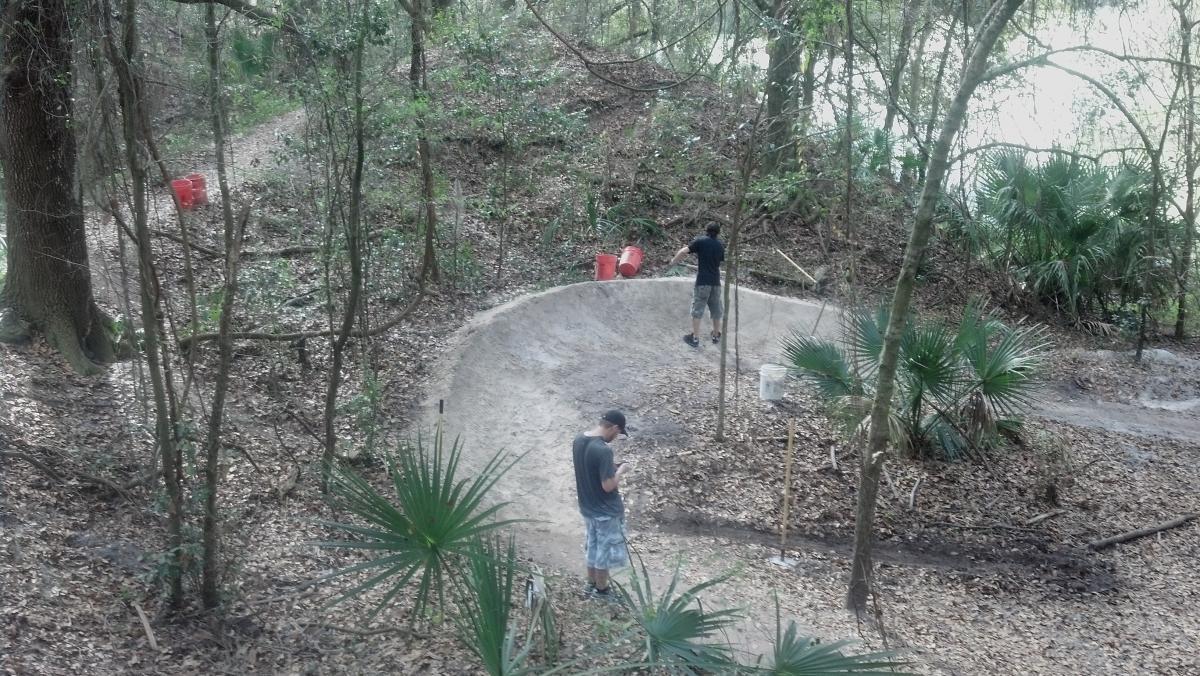 Two individuals are working on a dirt trail in a wooded area. One person is using a shovel on a dirt embankment, while the other is standing nearby, checking a device. The background features trees, palm plants, and dirt paths, with bright red buckets visible among the foliage. Alafia River State Park mountain bike trail.