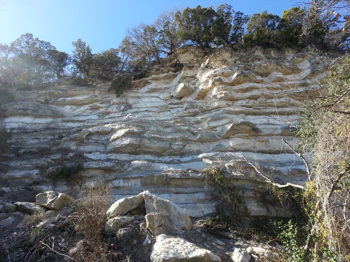 A steep, layered rock formation with visible striations and vegetation along the top, under a clear blue sky. The foreground shows scattered rocks and dry brush, leading up to the rugged cliff face. Cameron Park mountain bike trail.