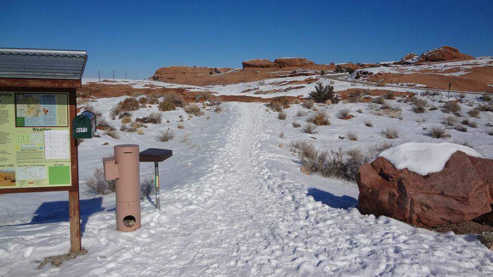 A snow-covered pathway leads through a winter landscape, with reddish rock formations in the background. On the left, a informational sign outlines weather and trail details, and a green mailbox is attached nearby. A brown waste disposal unit stands beside the path, which is clearly marked with footstep impressions from previous visitors. The sky above is clear and blue. Slickrock mountain bike trail.