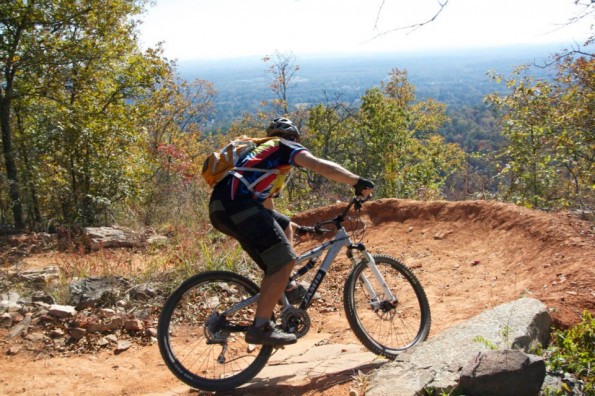 A mountain biker navigating a dirt trail downhill through a forested area, with colorful autumn foliage and a distant view of rolling hills in the background. The cyclist is wearing a helmet and a multi-colored jersey, and has a backpack.