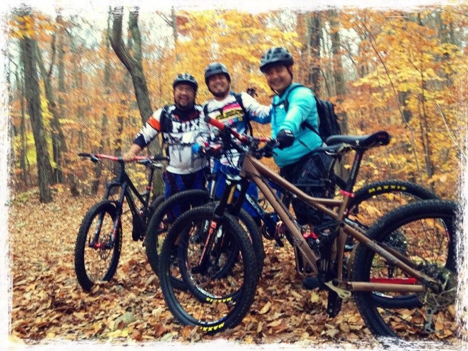 Three mountain bikers pose for a photo in a wooded area during autumn. They are surrounded by trees with colorful fall foliage and a carpet of leaves on the ground. Each rider is wearing biking gear and holding their bikes, smiling at the camera. Ringwood Skylands Manor mountain bike trail.