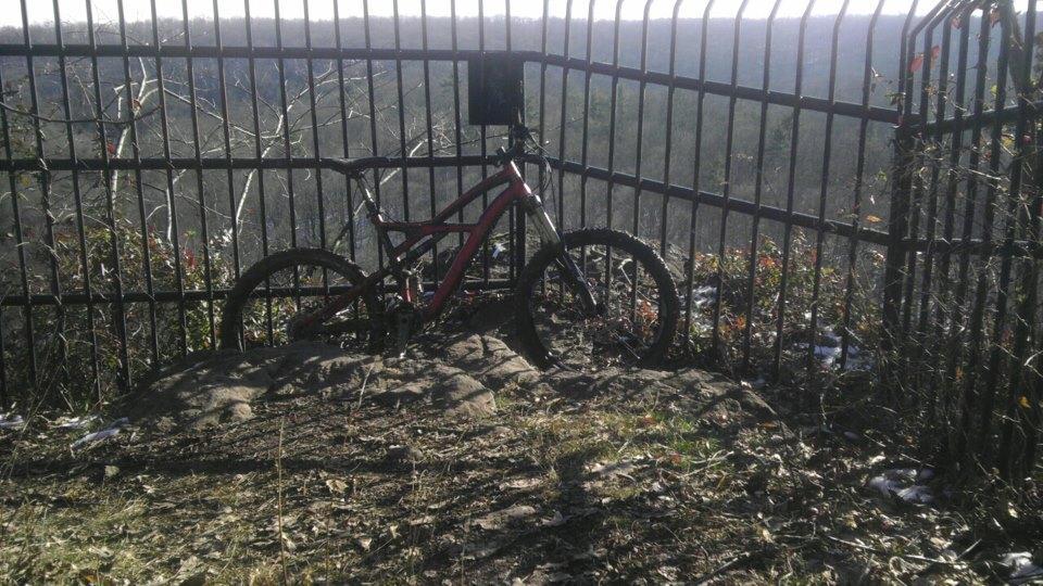 A red mountain bike is placed against a rocky surface inside a fenced area overlooking a wooded landscape. The background features trees and a distant view, with some vegetation and grass in the foreground. The scene is illuminated by daylight, highlighting the bike and natural surroundings. High Rocks mountain bike trail.