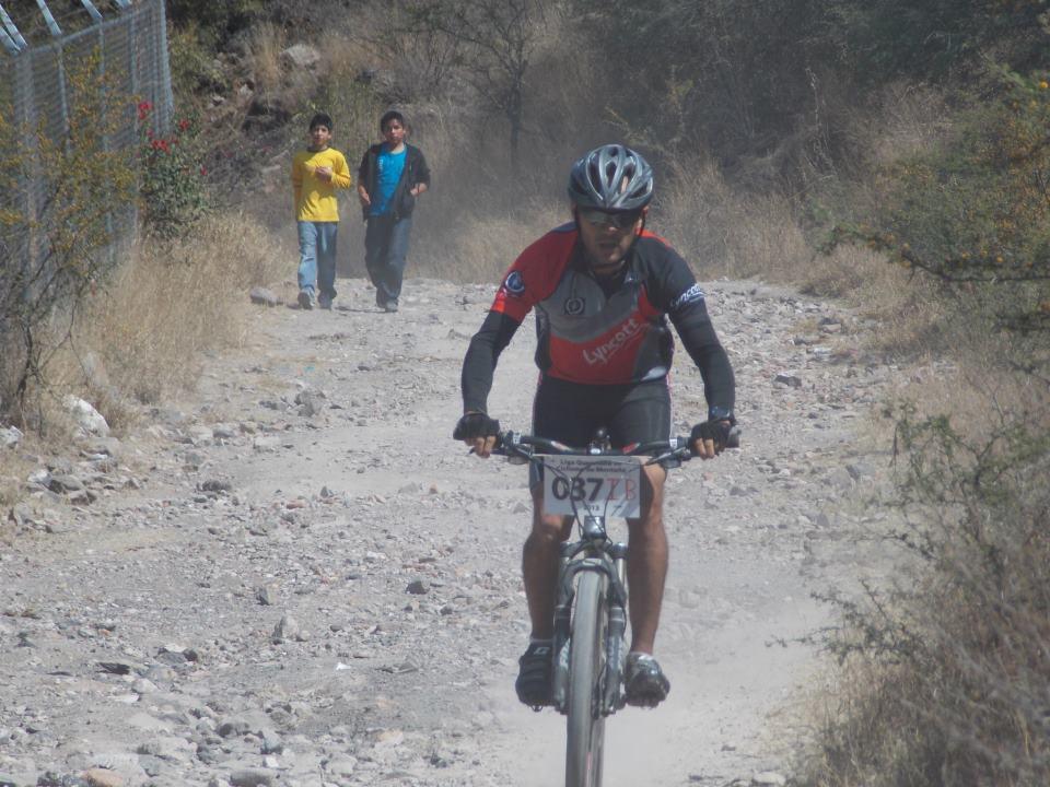 A mountain biker rides along a rocky dirt path, kicking up dust in a rural setting. In the background, two young boys walk casually on the same path, one wearing a yellow shirt and the other in a blue shirt. Foliage and a fence are visible on the left side of the image. Santa Barbara 2013 mountain bike trail.