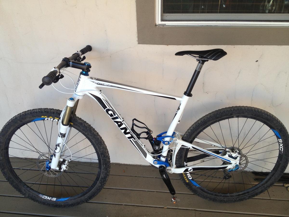 Giant Anthem X 29er 3: A white mountain bike with black and blue accents leaning against a wall. The bike features a suspension fork, knobby tires, and ergonomic handlebars. The background shows a light-colored wall and a window, with the bike resting on a wooden deck.