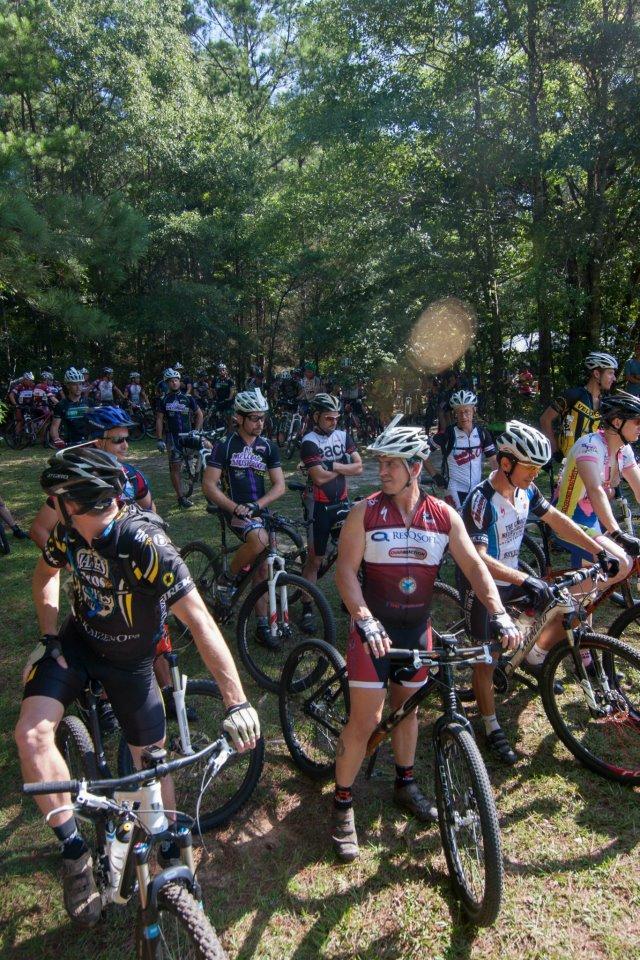 A large group of mountain bikers in cycling attire, wearing helmets, gathered in a wooded area. The scene captures both men and women standing near their bikes, surrounded by trees and greenery, with a sense of camaraderie and anticipation for an upcoming race or event. Chewacla State Park mountain bike trail.