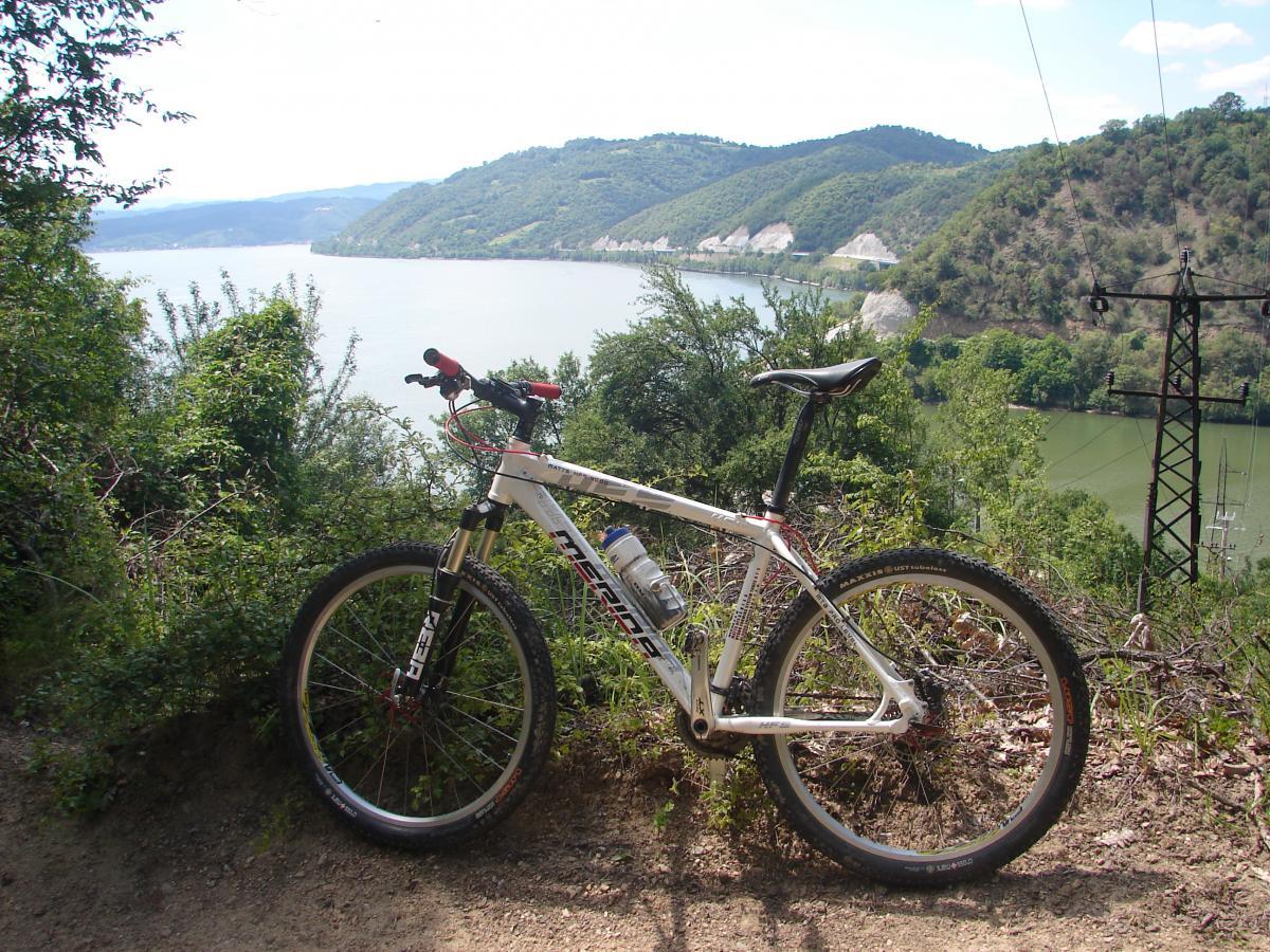 Merida Merida HFS: A mountain bike parked on a dirt path with a scenic view of a river and hills in the background. The surrounding area is lush with greenery and trees under a bright sky.