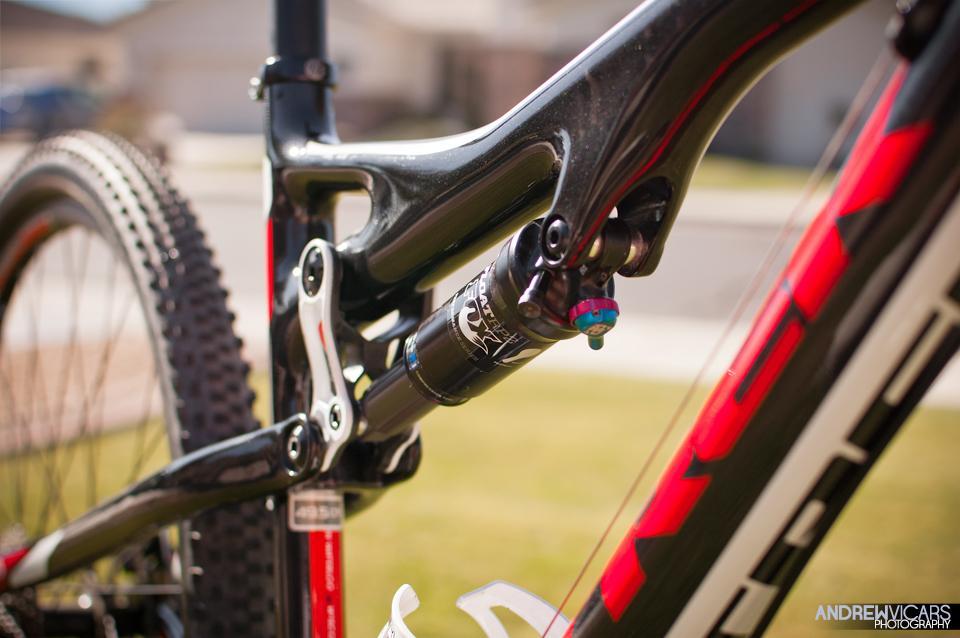 Trek Superfly Full Suspension: Close-up view of a mountain bike's rear suspension system, showcasing the shock absorber and frame details. The bike features a black and red color scheme, set against a blurred outdoor background with grass and a house.
