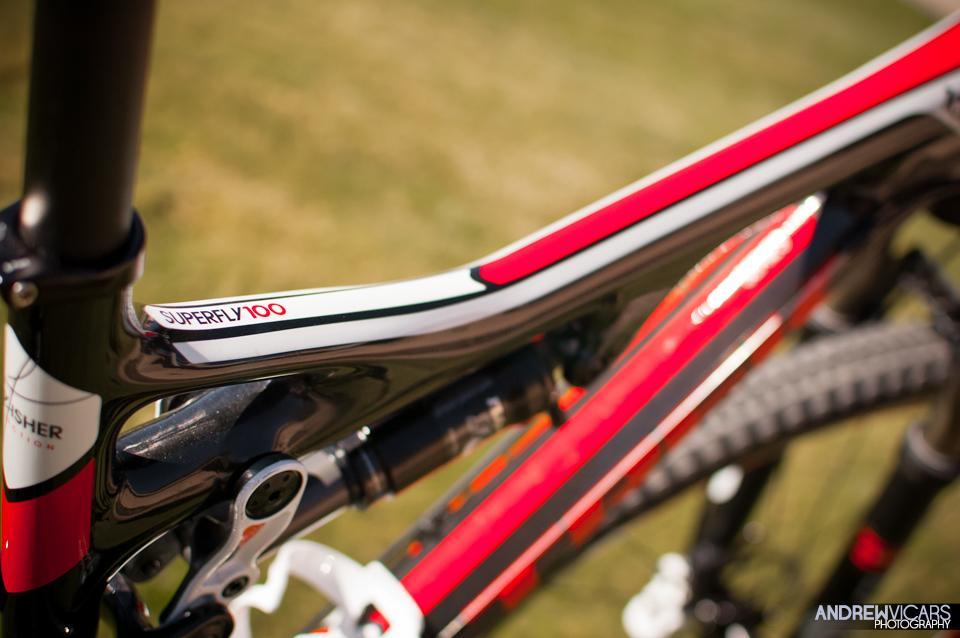 Trek Superfly Full Suspension: Close-up image of a bike frame featuring the "Superfly 100" branding in a bold design, with vibrant red and black colors, highlighting the sleek contours and intricate details of the bike's construction. The background is an out-of-focus grassy area.