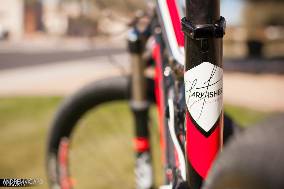 Trek Superfly Full Suspension: A close-up view of a red and white bicycle frame featuring the "Gary Fisher Collection" logo on the down tube, with a blurred background showcasing grass and a pathway.
