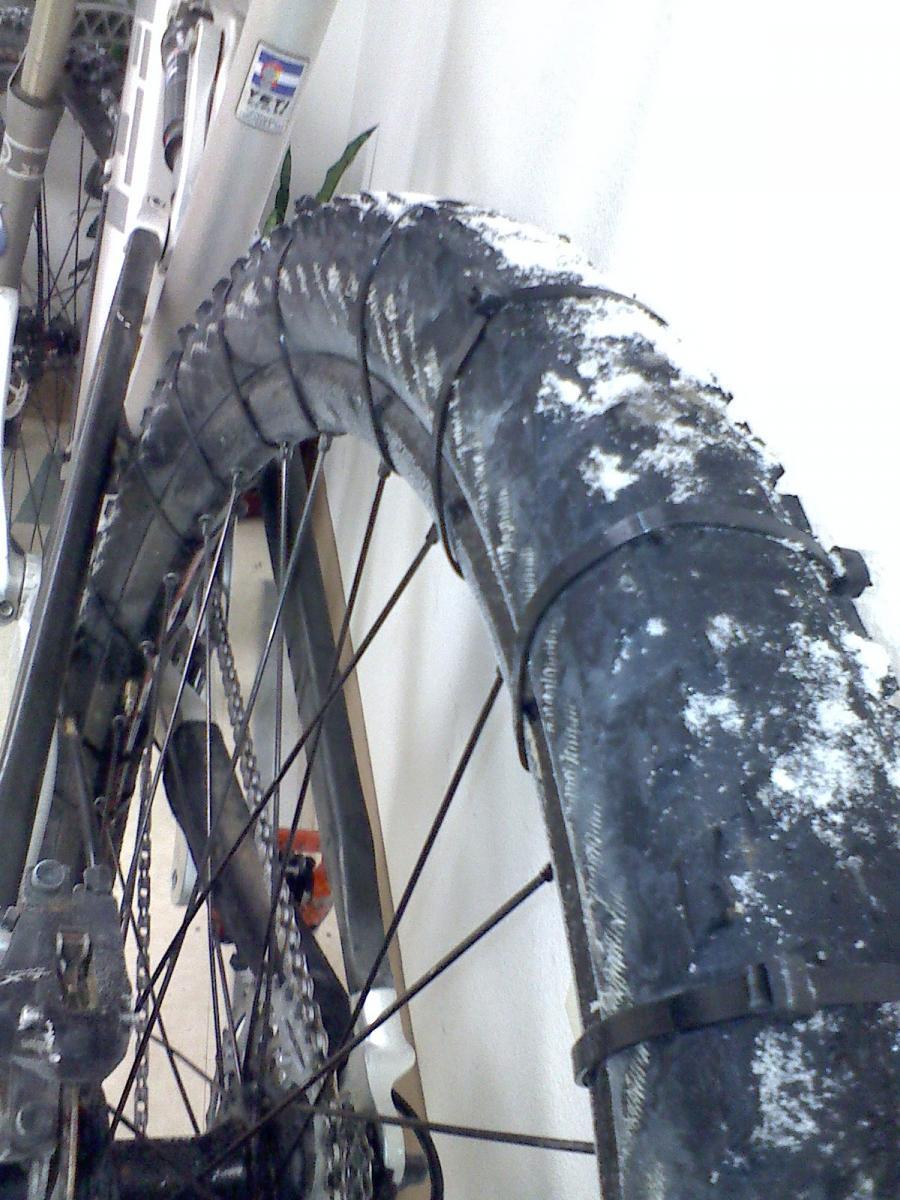 A close-up view of a mountain bike tire, partially covered in snow, mounted on a bike frame. The tire's tread pattern is visible, along with the spokes and chain of the bike. A light-colored wall and a plant can be seen in the background. Slickrock mountain bike trail.