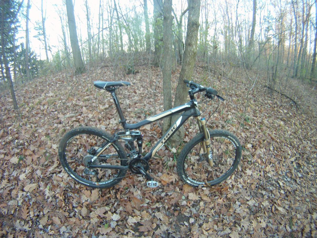 Trek Fuel EX 7: Mountain bike resting on a bed of fallen leaves in a wooded area, with trees in the background. The bike features a sleek black and silver design and is positioned on a slight incline.