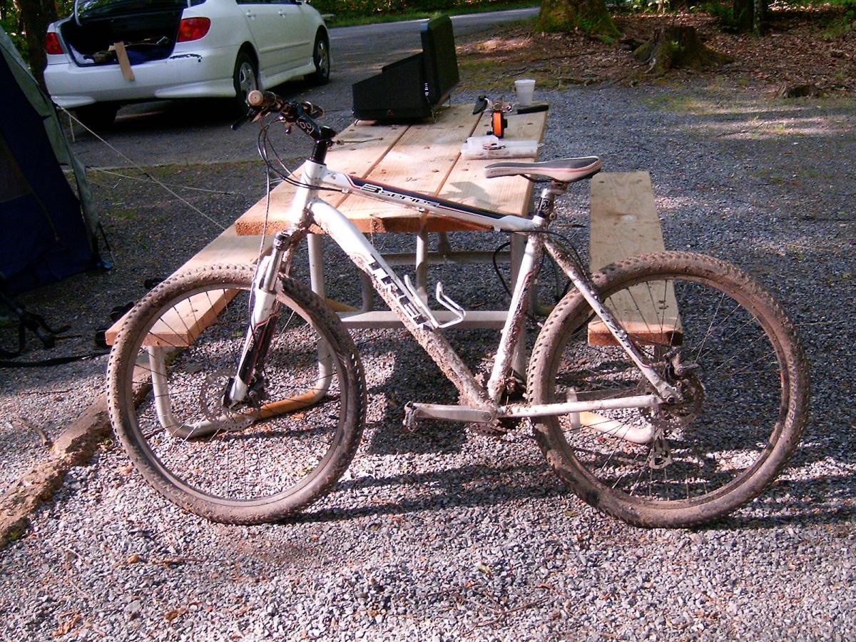 Trek 3700: A mountain bike with a white frame and muddy tires is parked beside a wooden picnic table in a gravel campsite. In the background, a car is partially visible, with its trunk open, among a forested area.