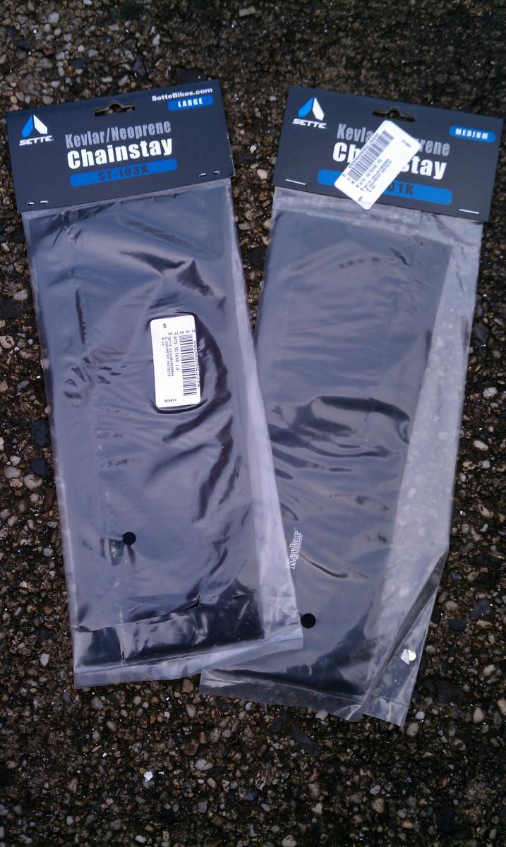 Sette Kevlar/Neoprene Chain Stay Protector: Two packages of Kevlar/Neoprene chainstays from Sette, one labeled 'Large' and the other 'Medium,' displayed on a textured surface. Each package is clear, revealing the black chainstays inside and includes product labels with barcodes.
