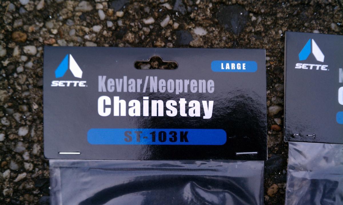 Sette Kevlar/Neoprene Chain Stay Protector: Black packaging for a chainstay made of Kevlar and neoprene, labeled as "Sette," with "Large" and "ST-103K" clearly visible. The background shows a textured surface of gravel.