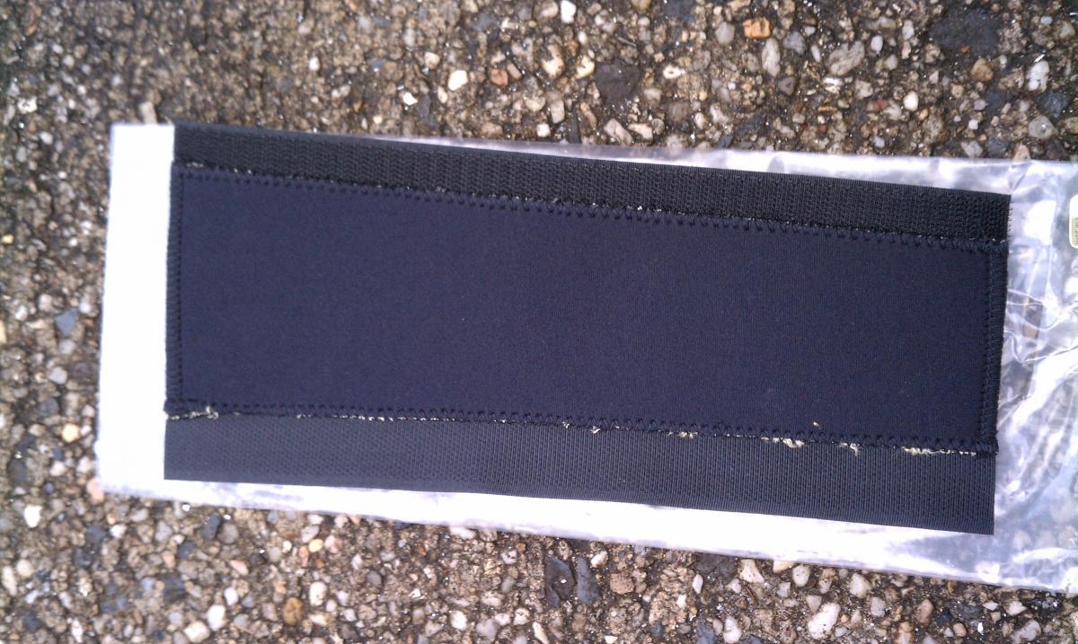 Sette Kevlar/Neoprene Chain Stay Protector: Black fabric patch with stitched edges, placed on a clear plastic wrapper, resting on a textured asphalt surface.