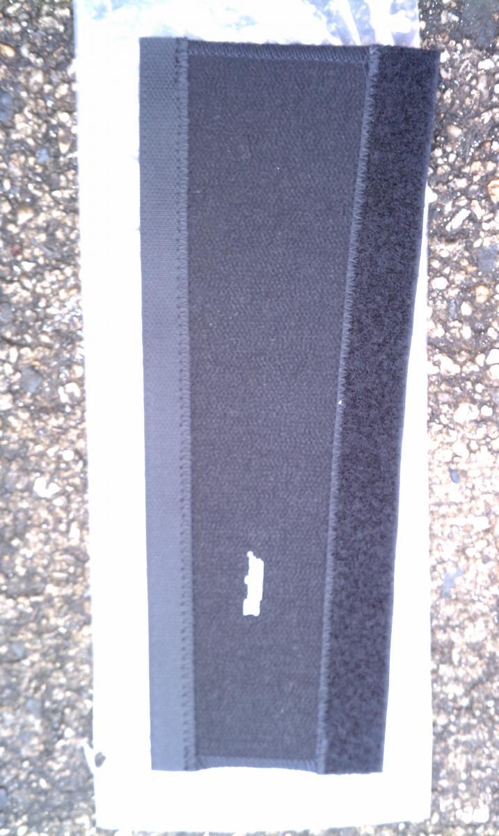 Sette Kevlar/Neoprene Chain Stay Protector: A close-up image of a rectangular fabric piece featuring various textures and colors, primarily black with gray and velcro-like sections. The fabric is laid out flat on a textured surface, possibly concrete, with a hint of a plastic wrapper visible at the edges.