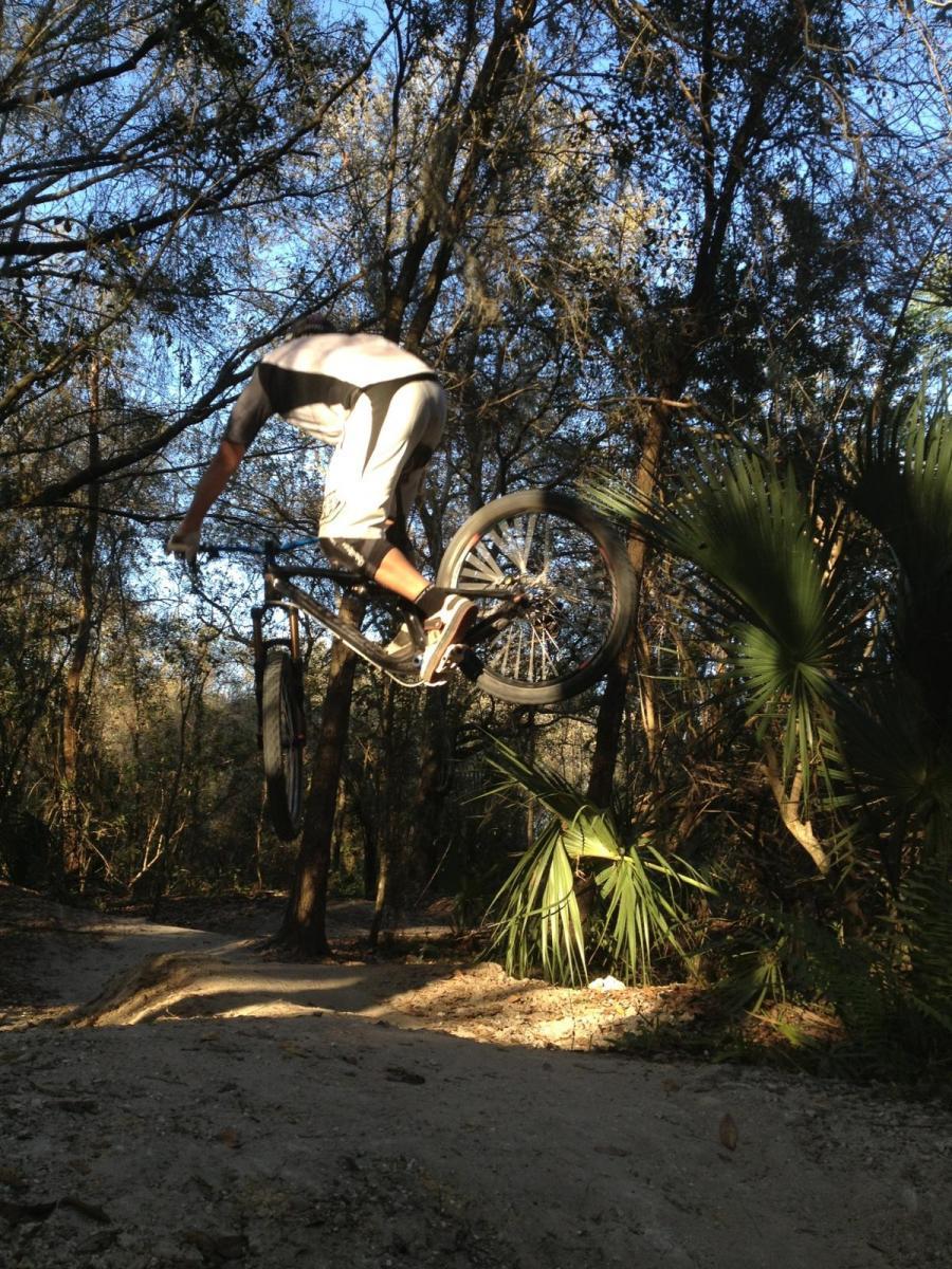 A person performing a jump on a mountain bike in a forested area, surrounded by trees and palm plants. The rider is captured mid-air, showcasing an action shot against a backdrop of blue sky and natural foliage. Alafia River State Park mountain bike trail.