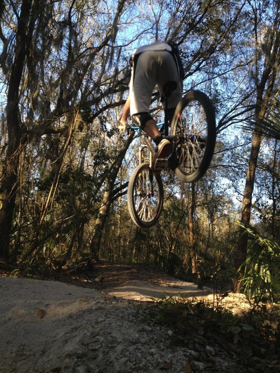 A person jumping off a dirt ramp on a mountain bike, surrounded by trees in a forested area. The rider is airborne with their bike, showcasing a dynamic mid-air stunt against a clear blue sky. Alafia River State Park mountain bike trail.