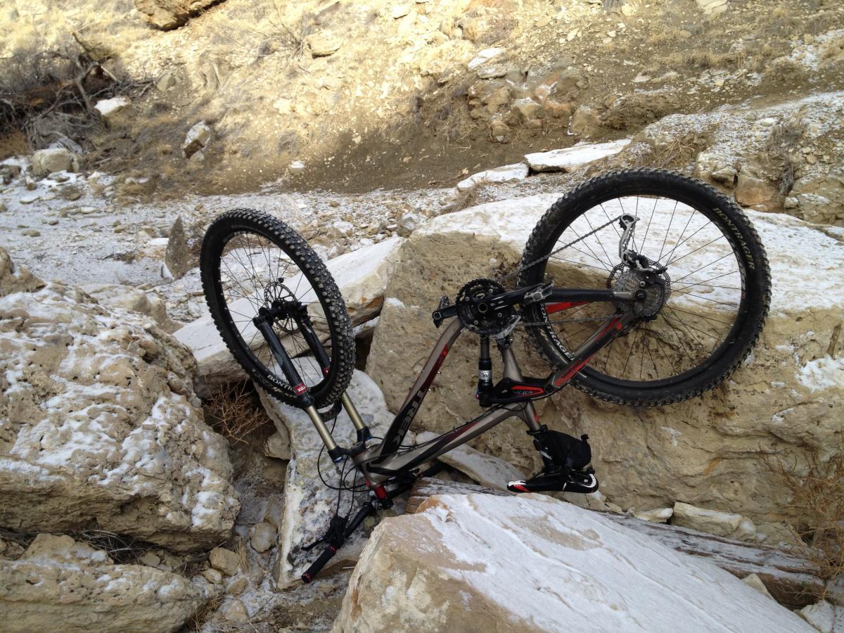 A mountain bike lying upside down on rocky terrain, with its front wheel resting against large boulders and a gravelly path in the background. The scene suggests a rugged outdoor environment, possibly an off-road trail. South Shore Lake Pueblo mountain bike trail.