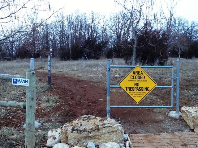 A gate with a yellow "Area Closed - No Trespassing" sign stands at the entrance of a rural area. The surrounding landscape features a grassy field and sparse trees in the background. A wooden fence is visible on the left with a sign that reads "MANN." The scene appears to be overcast, indicating a cool or early evening setting. Shawnee Mission Park mountain bike trail.