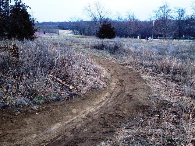 A winding dirt path through a grassy area, bordered by sparse foliage and bare trees, leading into the distance. The sky is overcast, creating a muted atmosphere. Shawnee Mission Park mountain bike trail.