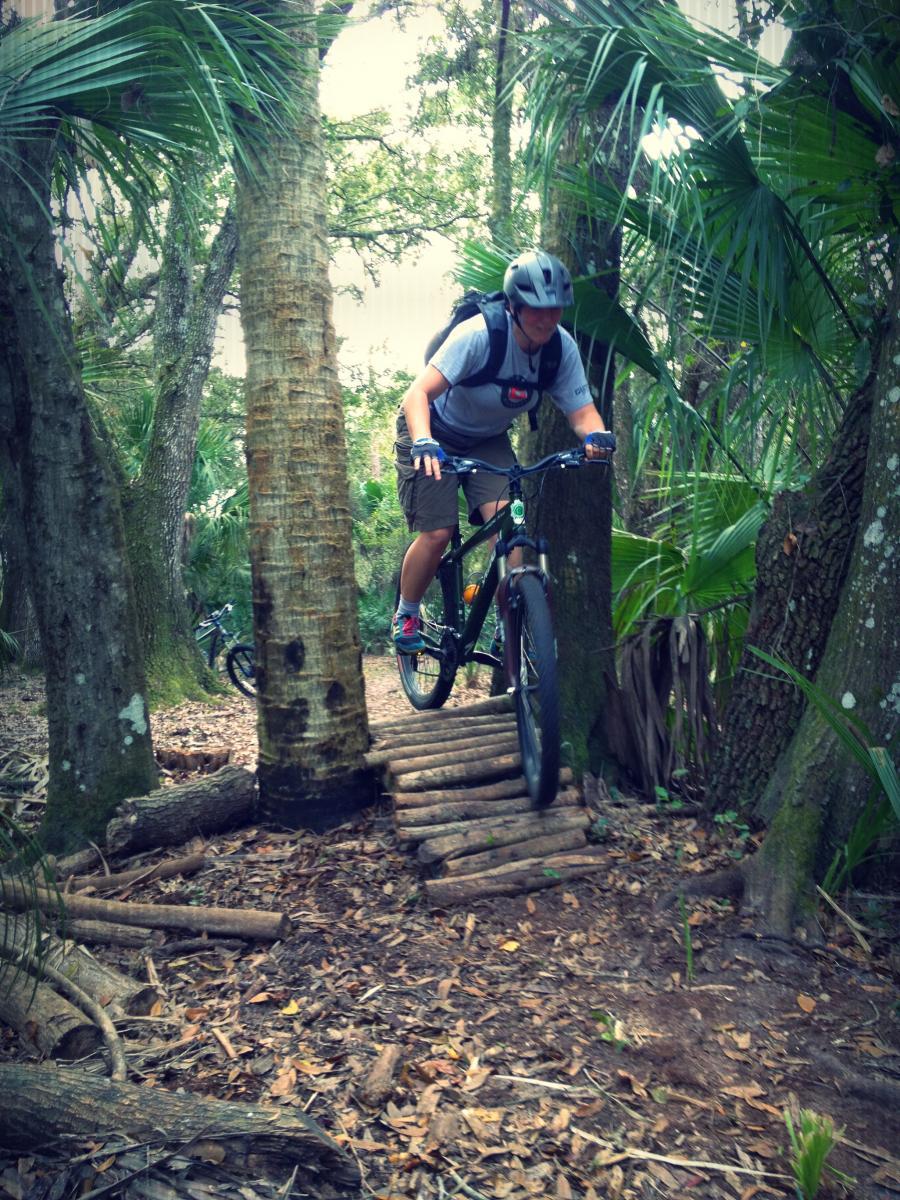 A mountain biker maneuvering over a wooden bridge in a lush, wooded area filled with palm trees and foliage. The cyclist wears a helmet and riding gear, focused on navigating the trail ahead. Grassy Island Trail mountain bike trail.