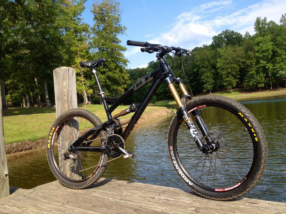 Yeti SB66: A black mountain bike is leaning against a wooden post on a dock, with a calm pond in the background. The bike features large tires with visible tread patterns, and the setting includes lush green trees and a blue sky.