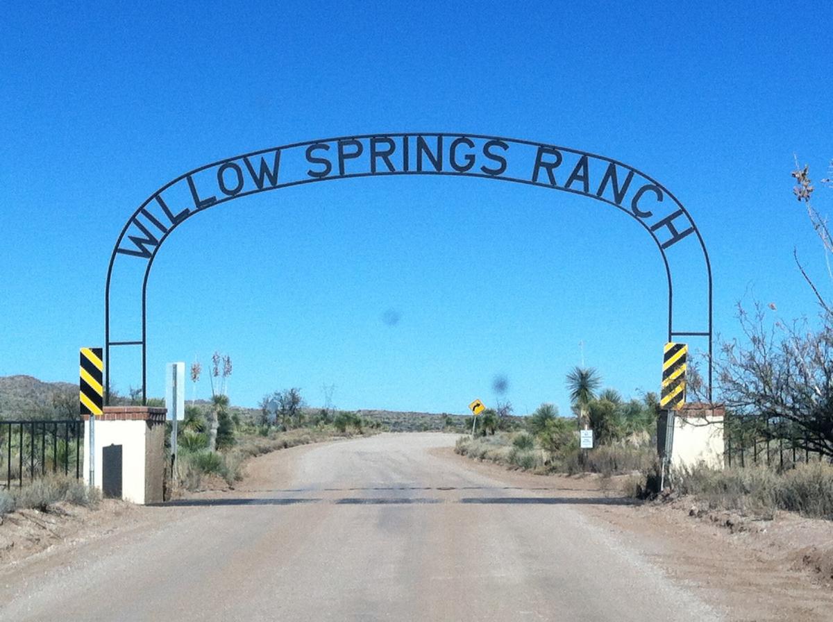 Image of the entrance gate to Willow Springs Ranch, featuring an archway sign with the text "WILLOW SPRINGS RANCH" against a clear blue sky. The dirt road leads towards the ranch, with desert vegetation and sparse trees visible on either side. Black and yellow striped markers are positioned on the gate posts. 24 Hour Course mountain bike trail.