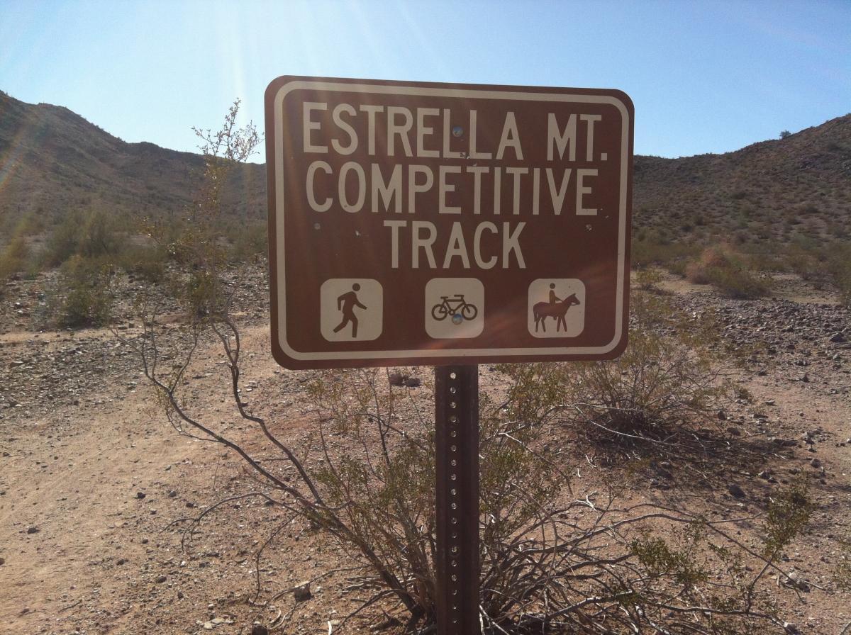 Sign for Estrella Mountain Competitive Track, indicating trail use for walking, biking, and horseback riding, against a backdrop of arid mountains and desert vegetation. Estrella Mountain Park mountain bike trail.