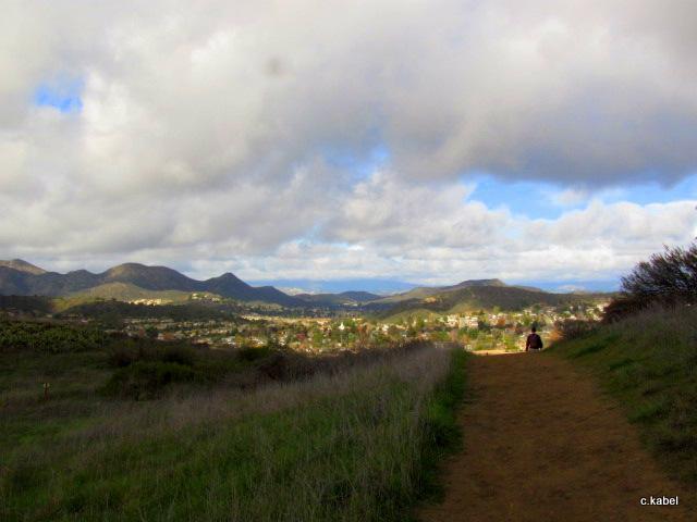 A scenic landscape featuring a winding dirt path leading through tall grass, with a person walking towards the horizon. In the background, rolling hills and mountains are visible beneath a partly cloudy sky, while a town sits nestled in the valley below. Los Robles Trail (Western Section) mountain bike trail.