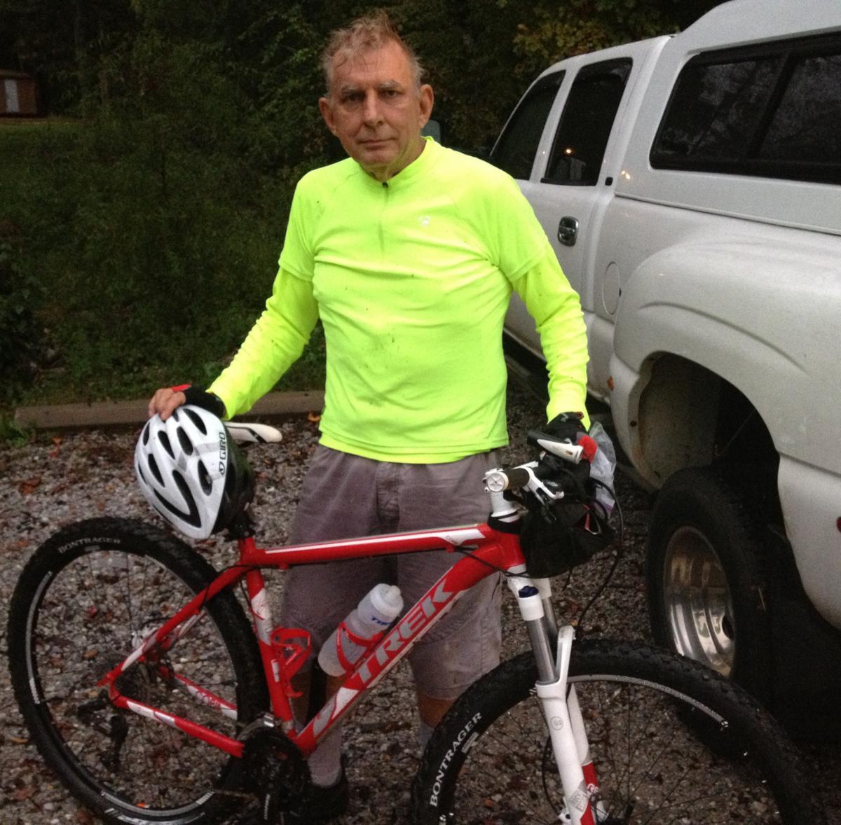 Trek Cobia: A man wearing a bright yellow long-sleeve shirt stands next to a red Trek mountain bike. He is holding a white helmet in one hand and the bike's handlebars with the other. The background features a white pickup truck and a gravel surface. The environment appears to be wooded, suggesting an outdoor setting.