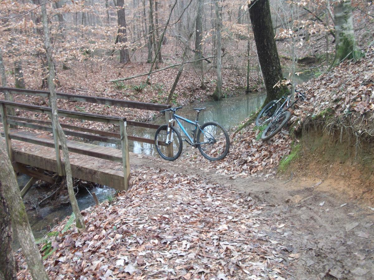 A peaceful forest scene featuring a wooden bridge over a small creek. Two bicycles are positioned near the bridge, with one resting on the ground and the other leaning against a tree. The ground is covered with fallen leaves, and surrounding trees have sparse foliage, indicative of a late autumn or early winter setting. Oak Mountain State Park mountain bike trail.