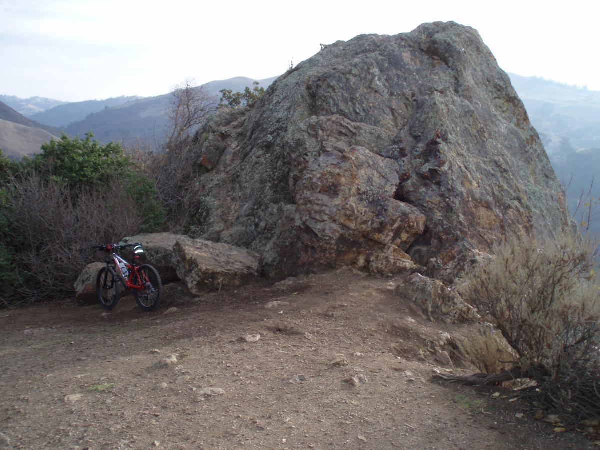 A mountain bike parked next to a large rock formation on a dirt trail, surrounded by sparse vegetation and rolling hills in the background under a cloudy sky. Alum Rock County Park mountain bike trail.