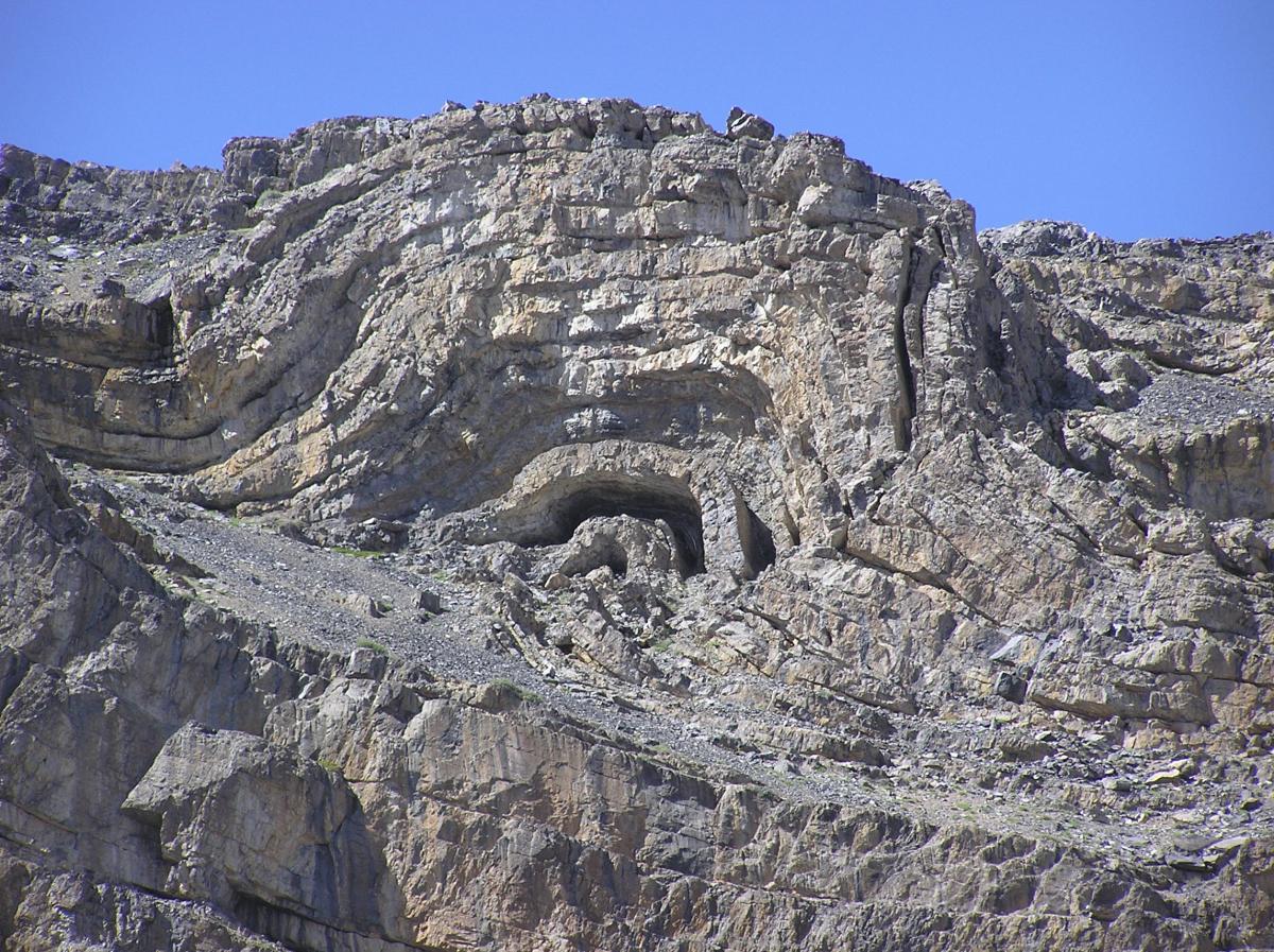 A rugged mountain landscape featuring layered rock formations, with a prominent cave opening partially visible in the rock face. The scene is set against a clear blue sky, highlighting the texture and contours of the rocky terrain. Webber Creek mountain bike trail.