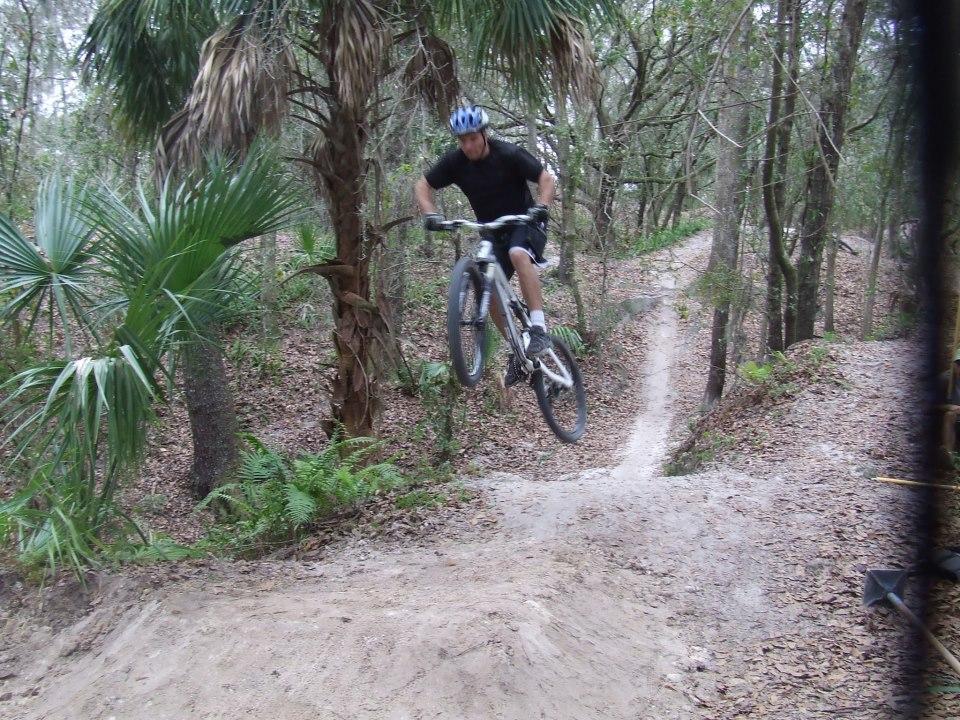 A person riding a mountain bike performs a jump on a dirt trail surrounded by trees and foliage. The rider is wearing a helmet and is airborne, with one hand on the handlebars while the bike's wheels are off the ground. The path curving in the background indicates the bike trail continues through the wooded area. Alafia River State Park mountain bike trail.