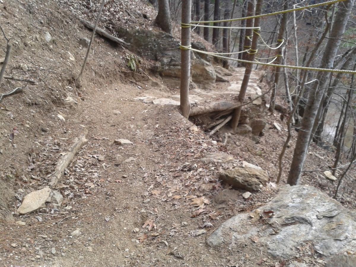 A narrow, dirt hiking trail winding through a wooded area, flanked by rocky outcrops and trees. A safety rope is anchored to nearby trees along the edge of the trail, which appears steep and has fallen leaves scattered on the ground. Van Michael Trail mountain bike trail.