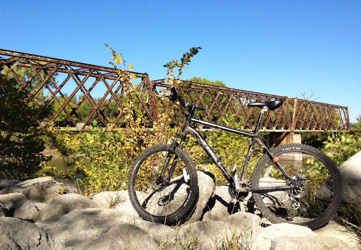 Trek 3700: A black mountain bike leaning against large rocks, with a rusty metal bridge in the background. The scene is set under a clear blue sky, surrounded by green foliage and hints of autumn colors.