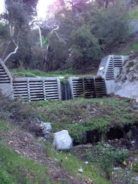 A concrete channel with a water flow spilling over slatted barriers, surrounded by greenery and trees in a natural setting. El Prieto mountain bike trail.
