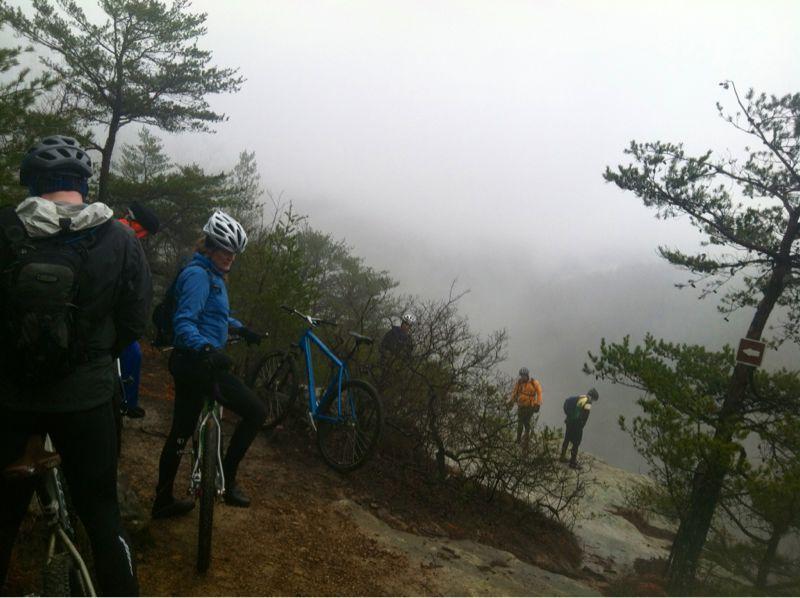 A group of mountain bikers pause on a rocky outcrop in a foggy landscape, with several bikes nearby. The scene is surrounded by trees, and the visibility is low due to mist, creating a serene and adventurous atmosphere. Some riders are looking out over the edge, while others chat and prepare for their ride. Big South Fork mountain bike trail.