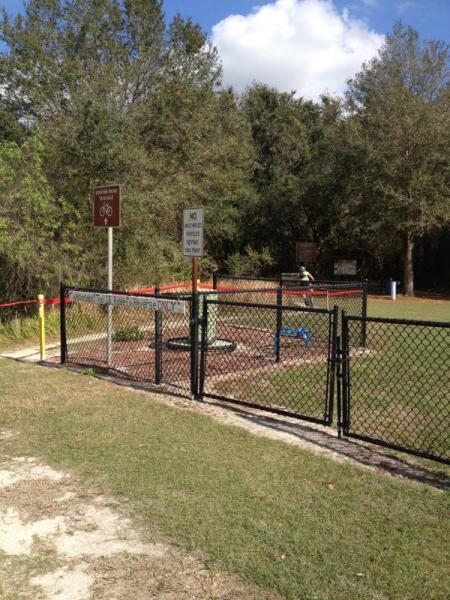 A fenced area with a green utility cover and a bicycle path sign nearby. The ground is grass, and there are trees in the background. A warning sign is visible, indicating prohibited actions, along with red tape marking off the area. Loyce E. Harpe Park mountain bike trail.