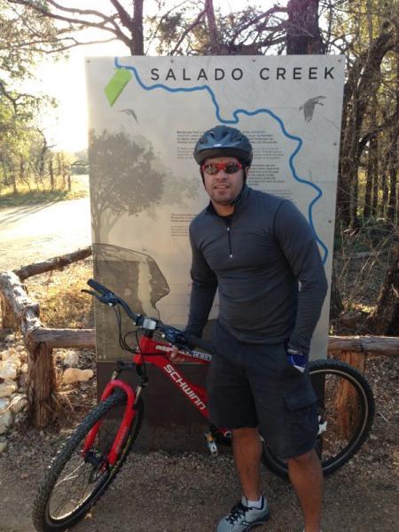 A person in cycling attire stands next to a trail sign indicating the Salado Creek path, holding a red Schwinn mountain bike. The background features trees and a dirt path, suggesting an outdoor recreational area. Salado Creek mountain bike trail.