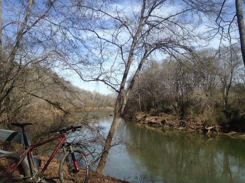 A scenic view of a calm river surrounded by bare trees and wooded areas, with a mountain bike resting on a wooden bench in the foreground. The sky is partly cloudy, adding a serene atmosphere to the natural landscape. Boundary Waters Park mountain bike trail.