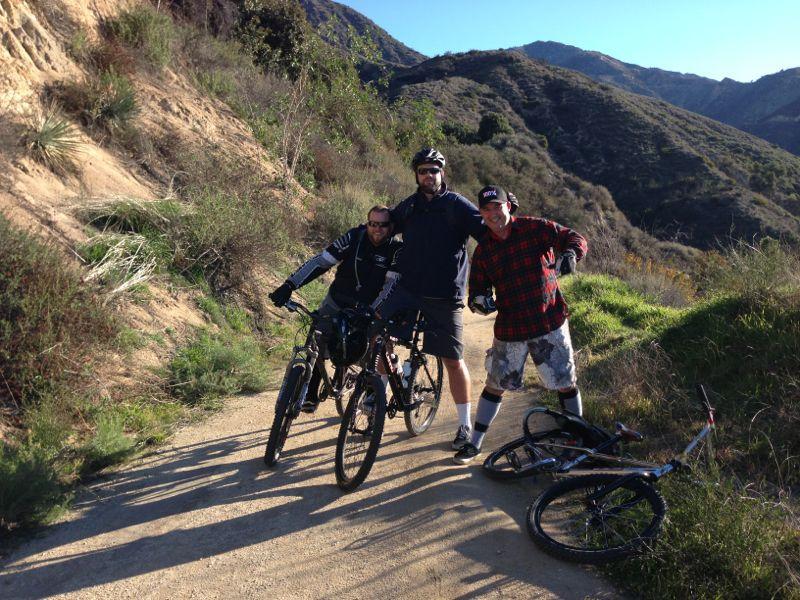 Three mountain bikers pose along a dirt path surrounded by greenery and hills. Two men stand next to their bikes, while one is crouching down. The setting is sunny with a clear blue sky, suggesting an outdoor adventure in a natural environment. One biker wears a plaid shirt, while the others are dressed in outdoor gear. One bike lies on the ground beside them. El Prieto mountain bike trail.