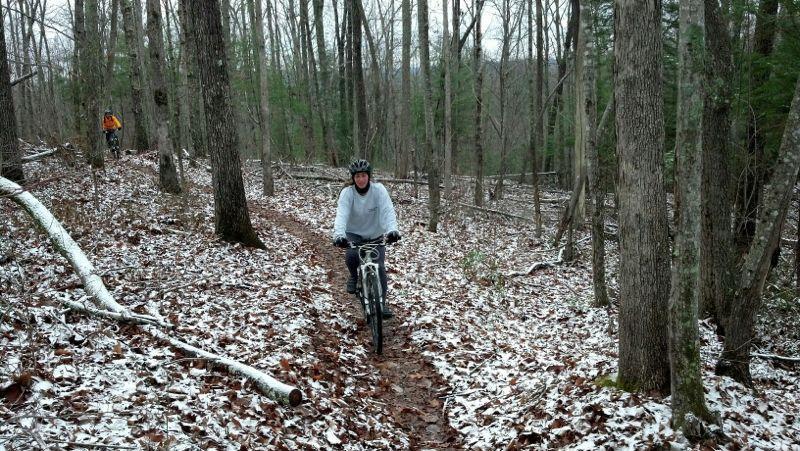 A cyclist wearing a helmet rides along a dirt path in a snowy forest, surrounded by tall trees and a carpet of fallen leaves. Another person in an orange jacket can be seen further back on the trail. Big South Fork mountain bike trail.