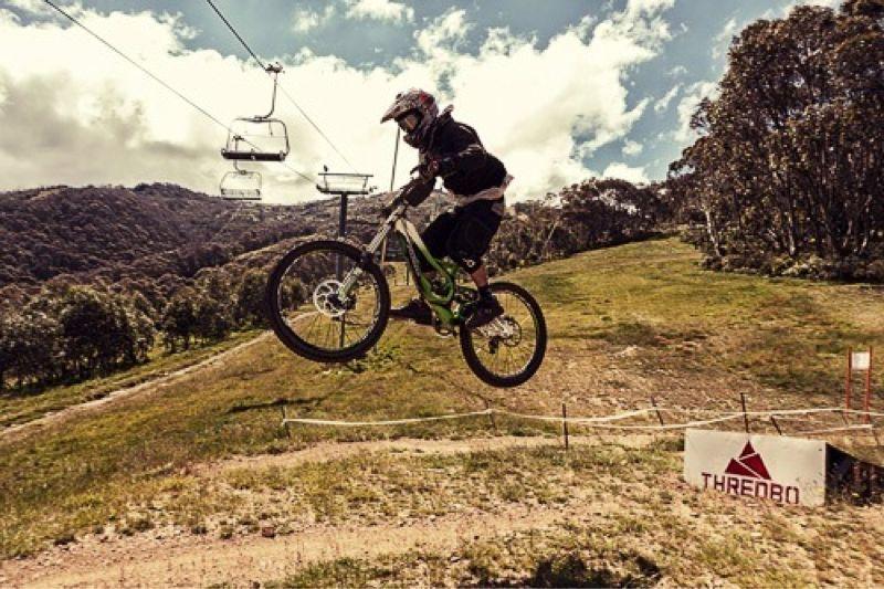 A mountain biker in protective gear jumps off a ramp on a green and black bicycle, soaring through the air above a grassy slope. In the background, a ski lift is visible under a partly cloudy sky, with trees and hills enhancing the outdoor setting. A sign for Thredbo is positioned nearby. Cannonball mountain bike trail.