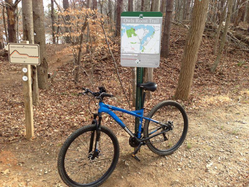 A mountain bike leans against a trail sign for "Owls Roost Trail" in a wooded area. The sign displays a map of the trail, and additional signage can be seen nearby. The ground is covered in brown leaves and gravel, with bare trees in the background, indicating an autumn or early winter setting. Owls Roost (Bur-Mil Park) mountain bike trail.