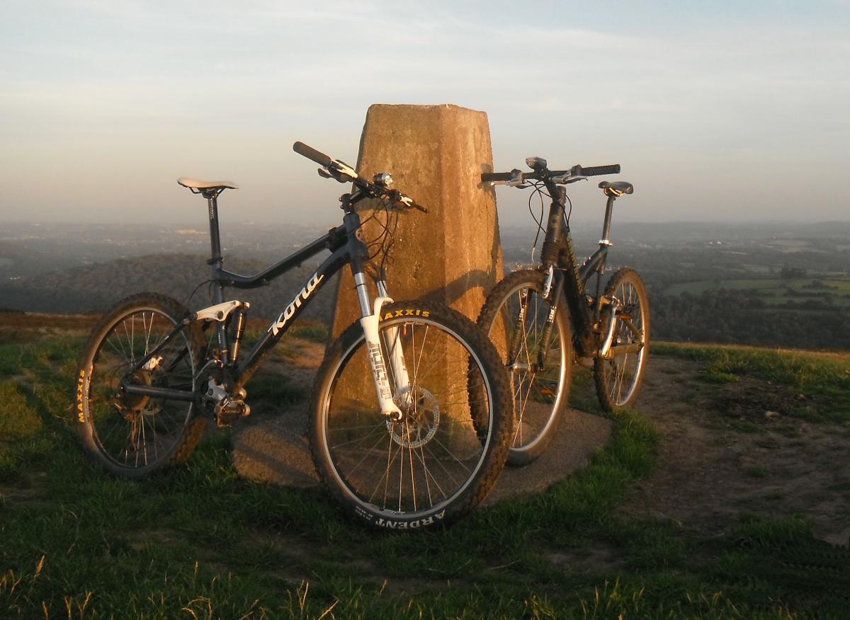 Kona Tanuki: Two mountain bikes are leaning against a stone marker on a hilltop during sunset. The landscape in the background features rolling hills and a clear sky. The bikes are black with distinct tire designs, showcasing a peaceful outdoor setting for cycling enthusiasts.
