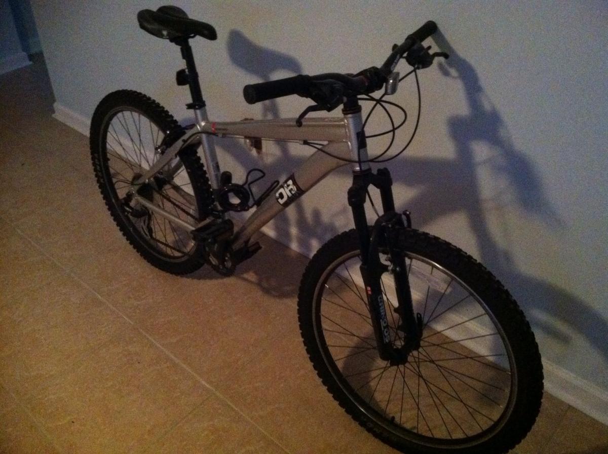 Diamondback Response: A grey mountain bike with thick, knobby tires is leaning against a wall on a tiled floor. The bike features a dual-suspension frame, black handlebars, and a padded seat. The setting is indoors, with soft lighting casting shadows on the wall.